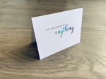 A white folded notecard is propped up on a wooden floor. It reads, "you are capable of anything." The word, anything, is in a script font that fades from green to navy. From Stationare's Inspire Collection.