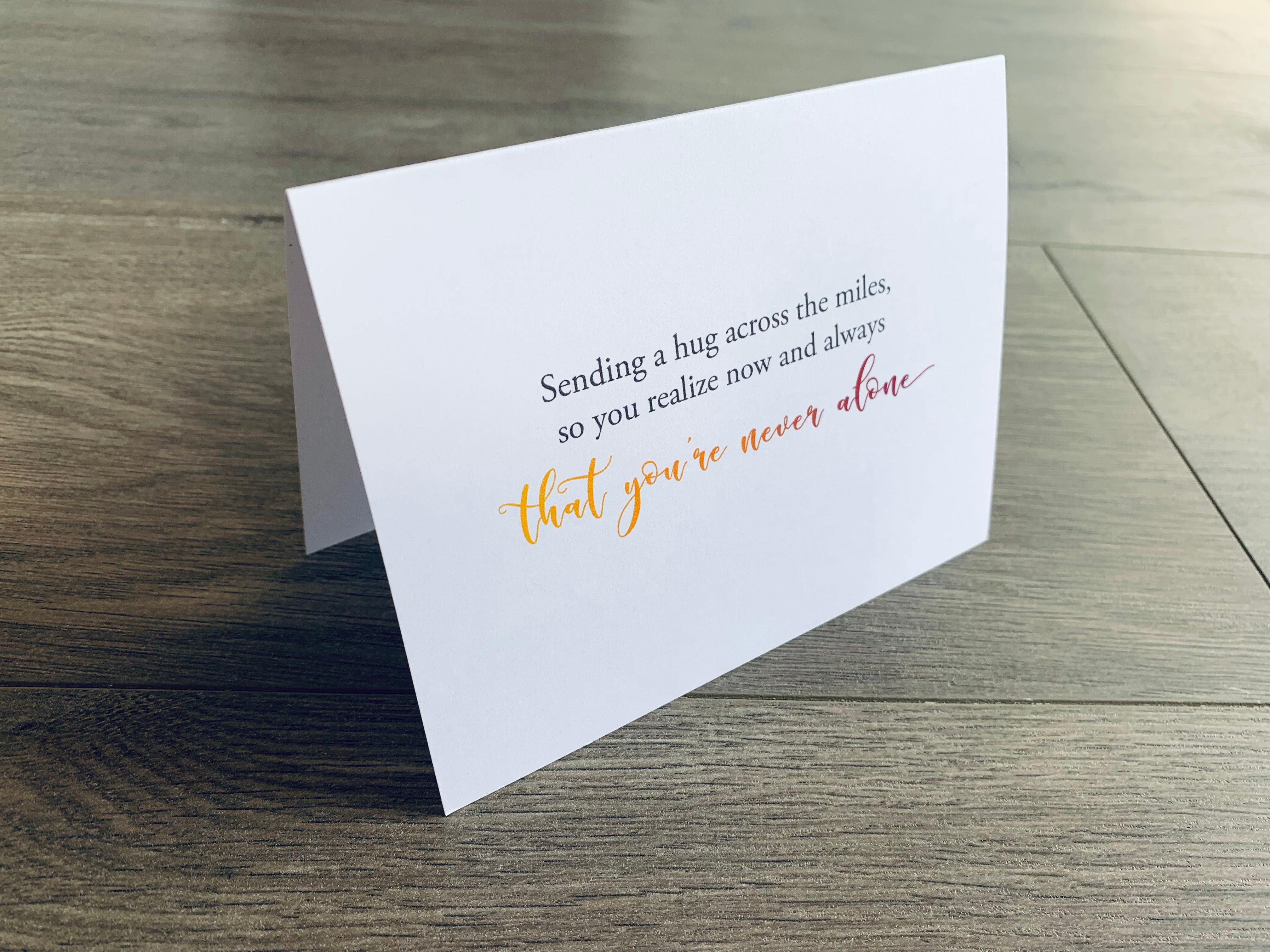 A white, folded notecard sits on a gray wooden floor. The card reads, "Sending a hug across the miles, so you realize now and always that you're never alone." Sending Support collection by Stationare.