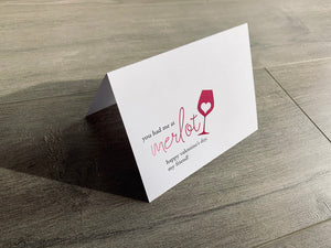 A folded white notecard is propped up on a wooden floor. The card says, "You had me at merlot. Happy Valentine's Day, my friend" with a heart inside a wine glass. Stationare's Valen-wine collection.